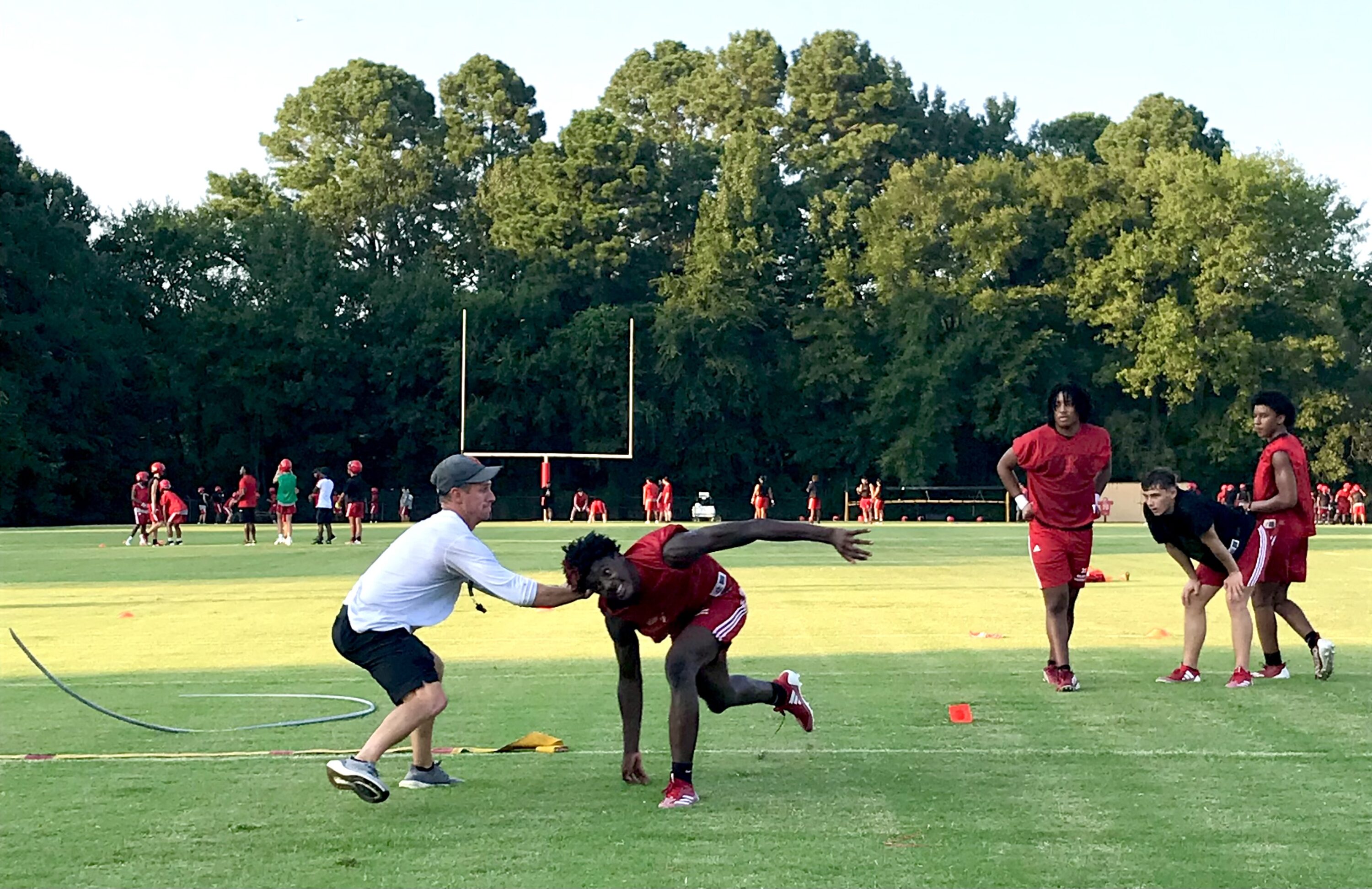 Kilgore High School head football coach Clint Fuller (left) works with outside linebackers on Day One: the first official day of UIL-approved high school football practice on Monday behind the Mitchell Field House. Below: the Ragin' Red meet briefly in the indoor practice facility. (Photos by JOE HALE)