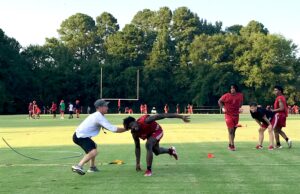 Kilgore High School head football coach Clint Fuller (left) works with outside linebackers on Day One: the first official day of UIL-approved high school football practice on Monday behind the Mitchell Field House. Below: the Ragin' Red meet briefly in the indoor practice facility. (Photos by JOE HALE)